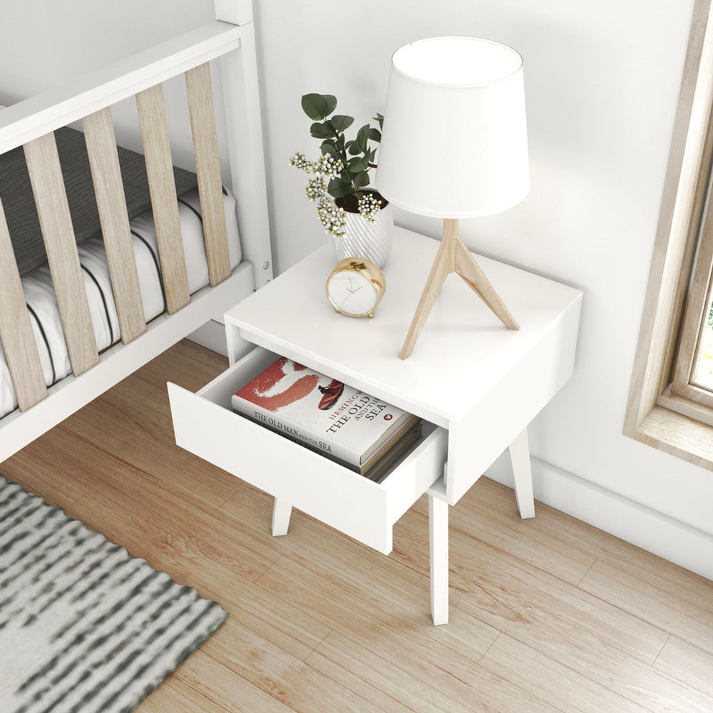 White modern nightstand with open drawer, wooden table lamp, gold clock, and vase with flowers next to crib in cozy bedroom setting.
