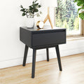 Matte black mid-century modern bedside nightstand with single drawer and tapered angled legs, styled with white vase, brass clock and wooden tripod lamp on light hardwood floor by a window