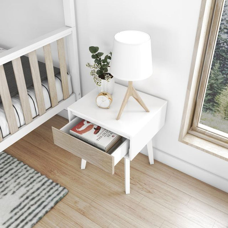 Scandinavian white bedside table with light-wood drawer, wooden tripod lamp, alarm clock and plant next to a baby crib on hardwood floor — minimalist nursery decor