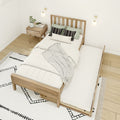 Scandinavian minimalist bedroom with wooden slat bed and pull-out trundle, white bedding and patterned pillows, wooden nightstand with plant and magazine, modern wall sconces, neutral boho rug