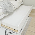 White wooden trundle bed with pull-out mattress, cream bedding and textured gray throw on light wood floor and patterned rug