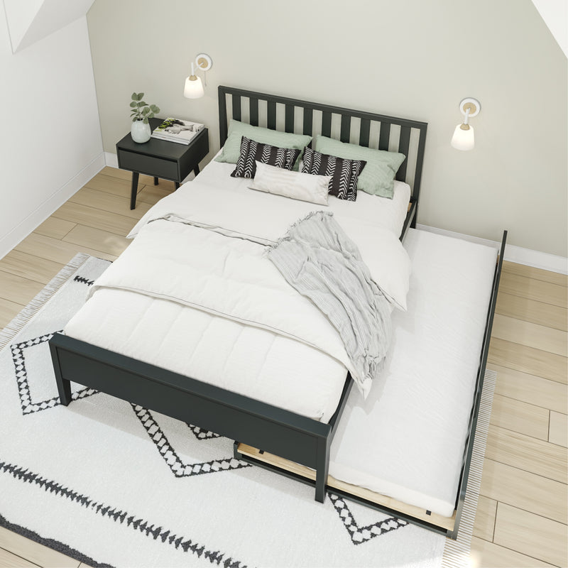 Minimalist bedroom with black wooden slatted bed frame and pull-out trundle, white bedding, green and black patterned pillows, black nightstand with plant and magazines, wall sconces, geometric rug on light hardwood floor