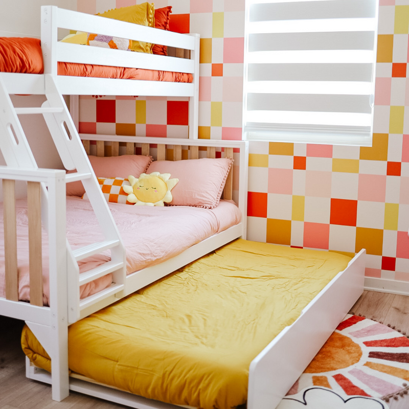 Colorful children's bedroom with a white bunk bed, pink bedding, and a cheerful sun pillow against vibrant checkered wallpaper.