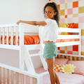 Smiling child climbing a white wooden bunk bed ladder in a colorful kids’ bedroom with orange bedding and a geometric accent wall