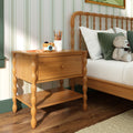 Natural wood nightstand with turned spindle legs and drawer beside bed with green pillows, teddy bear, toy camera and pencil cup