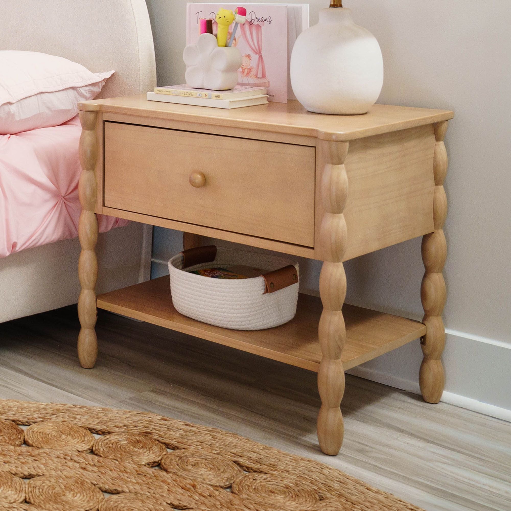 A dark-wood finish Jenny Lind style nightstand with one drawer and shelf next to a matching bed. There is a lamp and decor on the nightstand.
