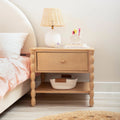 Light wood nightstand with single drawer, rattan table lamp, pink tufted bedding and woven storage basket in a cozy children's bedroom