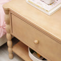 Light natural wood nightstand bedside table with single drawer, turned spindle legs and lower shelf with storage basket beside pink bed — children's bedroom nursery furniture