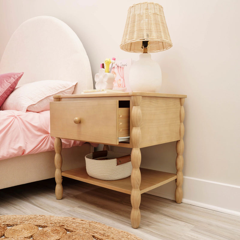 Light wood nightstand with turned legs and open drawer, wicker-shade table lamp, decorative accessories and woven basket beside pink bedding in a neutral bedroom