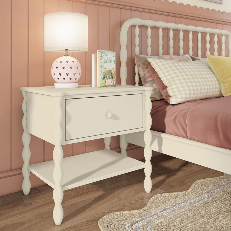 White scalloped bedside table nightstand with drawer and lower shelf next to pink cottage-style bed, heart-pattern pink lamp, gingham pillow and woven rug, cozy bedroom decor