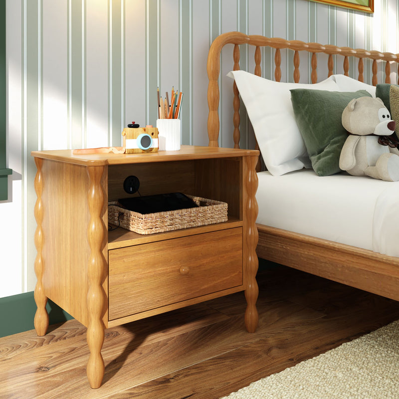 Wooden nightstand with turned legs in cozy kids bedroom — wicker basket with tablet, toy camera and pencils on top, adjacent wooden bed with green velvet pillows and plush teddy bear, striped wallpaper and hardwood floor
