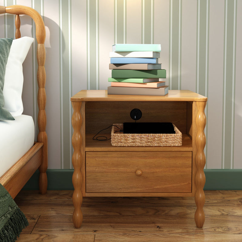 Wooden bedside table nightstand with turned legs, stack of pastel books on top, woven basket holding a charging tablet, next to a wooden bed and striped wallpaper in a cozy bedroom