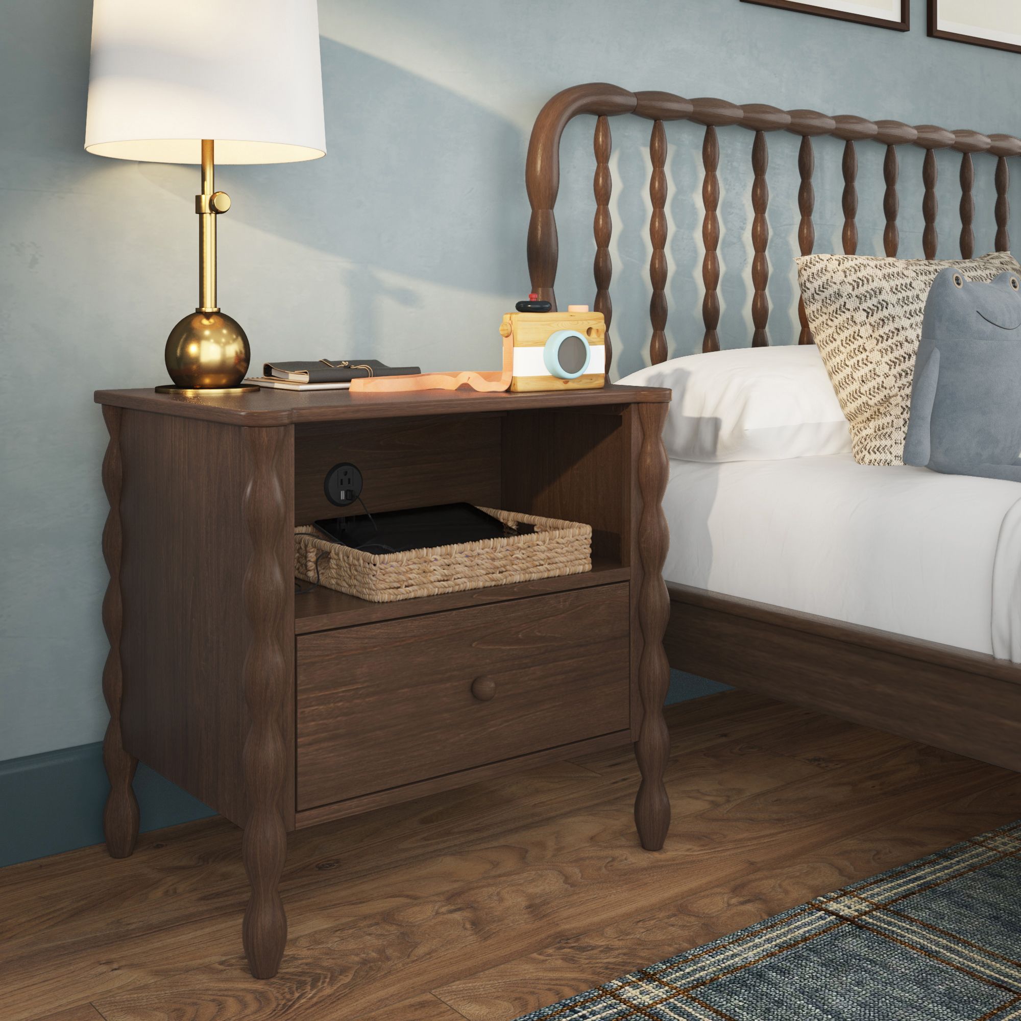 A light-wood finish Jenny Lind style nightstand next to a matching bed in a green room. The nightstand has a shelf with a charger port, holding a basket containing a device. On top of the nightstand is a lamp and decor.