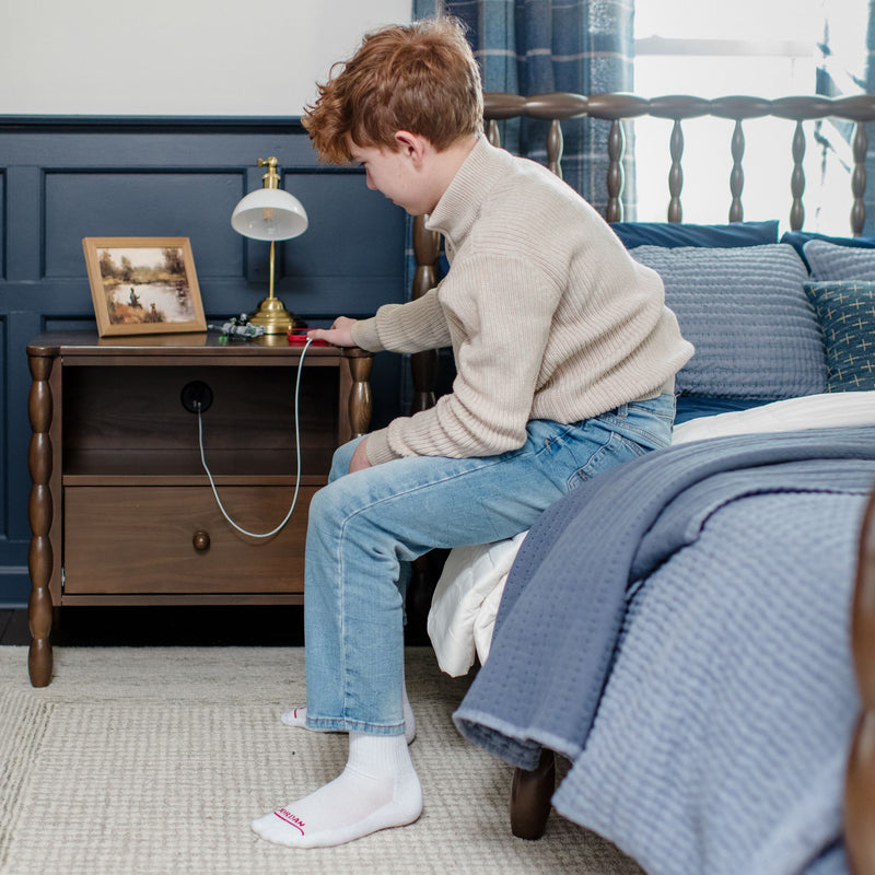 Boy plugging a phone charger into a bedside outlet on a wooden nightstand with lamp and blue bedding in a cozy bedroom.