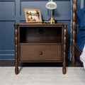 Brown wooden nightstand bedside table with turned spindle legs, open shelf with built-in power outlet, single drawer, brass lamp and framed art on top in a styled bedroom