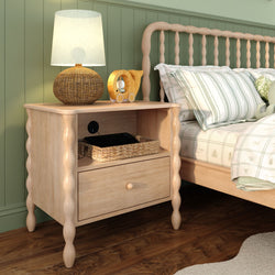 Light wood bedside table with drawer and shelf, rattan lamp, wicker basket and wooden toy elephant beside bed with striped pillows and green paneled wall