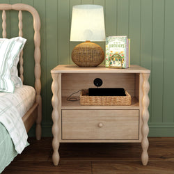 Light wood bedside table nightstand with turned spindle legs and drawer, topped with woven rattan lamp, wicker basket and gardening books against a green paneled wall in a cozy farmhouse bedroom