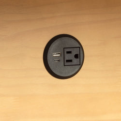 Round built-in desk power outlet with AC socket, USB-A and USB-C charging ports mounted in light wood surface