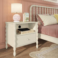 White spindle bedside table with drawer and open shelf holding a woven basket and charging tablet, pink heart-pattern lamp on top, gingham pillows and blush paneled wall in pastel bedroom decor