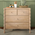Light wood nursery dresser against green paneled wall with woven basket holding plush frog, white pencil vase and wooden toy camera — children's room storage and decor