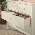 Cream painted wooden dresser with an open drawer revealing folded clothes, wicker basket, green plant and woven jute rug — bedroom storage furniture