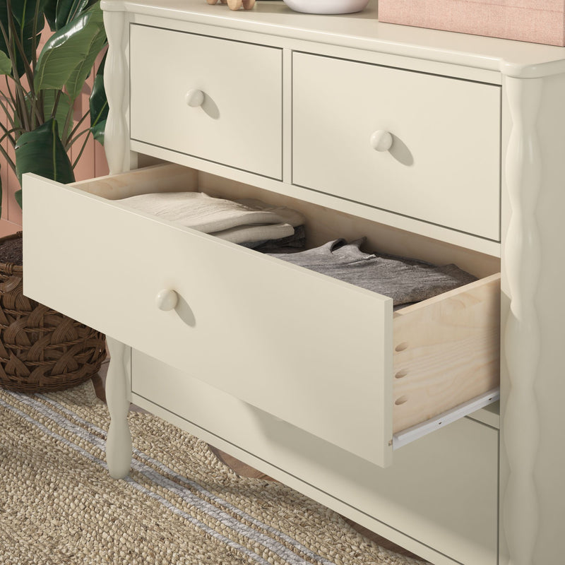 Cream painted wooden dresser with an open drawer revealing folded clothes, wicker basket, green plant and woven jute rug — bedroom storage furniture