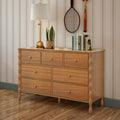 Light oak wooden dresser with eight drawers and turned legs in a cozy bedroom, topped with a gold table lamp, potted smiling planter, stacked books and vintage tennis racket against striped wallpaper