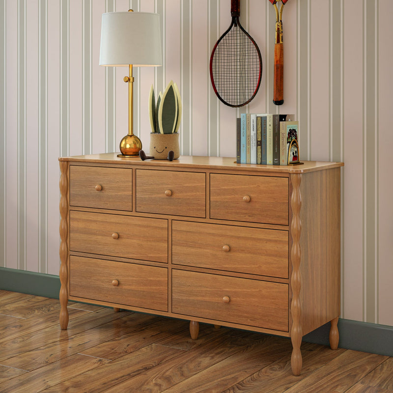 Light oak wooden dresser with eight drawers and turned legs in a cozy bedroom, topped with a gold table lamp, potted smiling planter, stacked books and vintage tennis racket against striped wallpaper