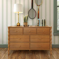 Wooden dresser with drawers on hardwood floor styled with brass table lamp, potted plant, stacked books and vintage tennis rackets against green striped wallpaper