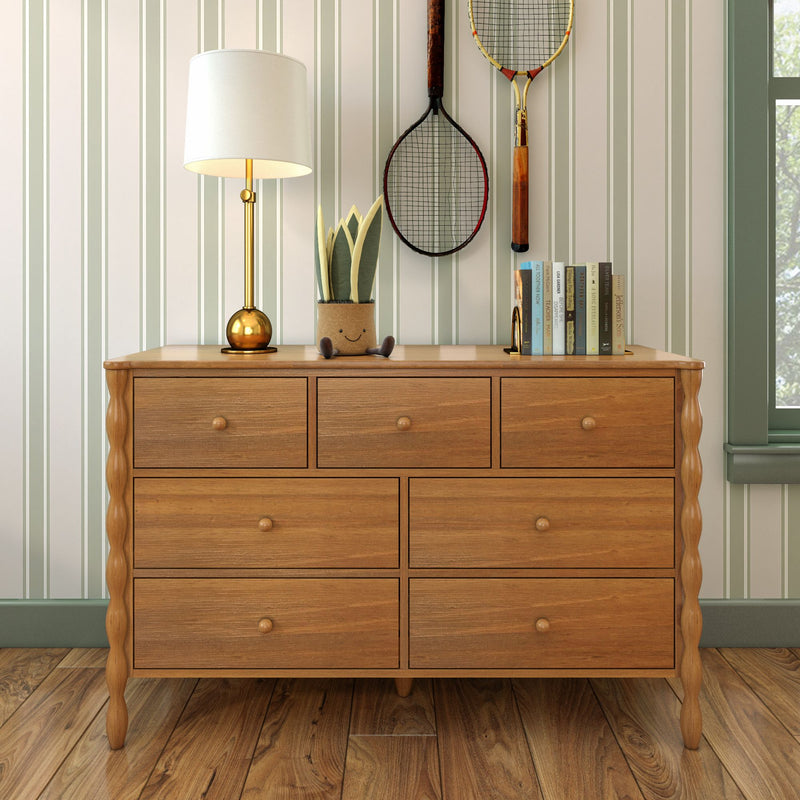 Wooden dresser with drawers on hardwood floor styled with brass table lamp, potted plant, stacked books and vintage tennis rackets against green striped wallpaper