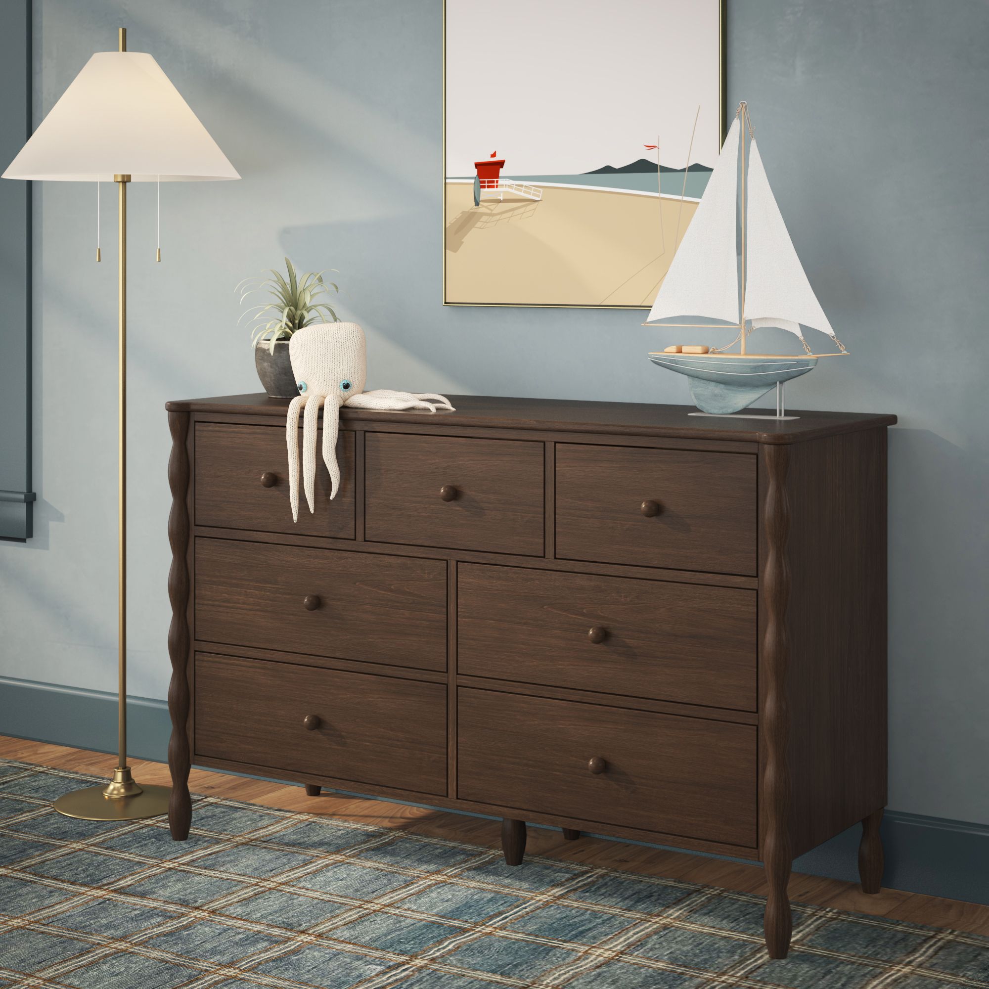 A light-wood Jenny Lind style 7-drawer dresser in a green room. A picture is hanging above the dresser, and small items including a pen cup and stuffed toy are on the dresser.
