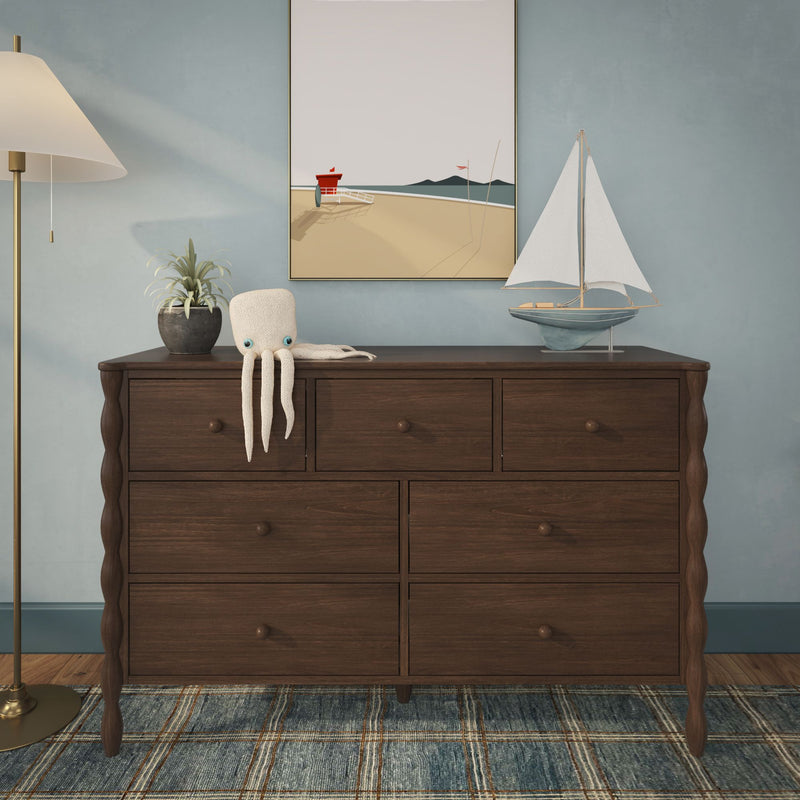 Dark wood six-drawer dresser with carved legs styled with nautical decor: model sailboat, knit octopus toy, potted plant, floor lamp and coastal beach print on a blue wall over a patterned rug