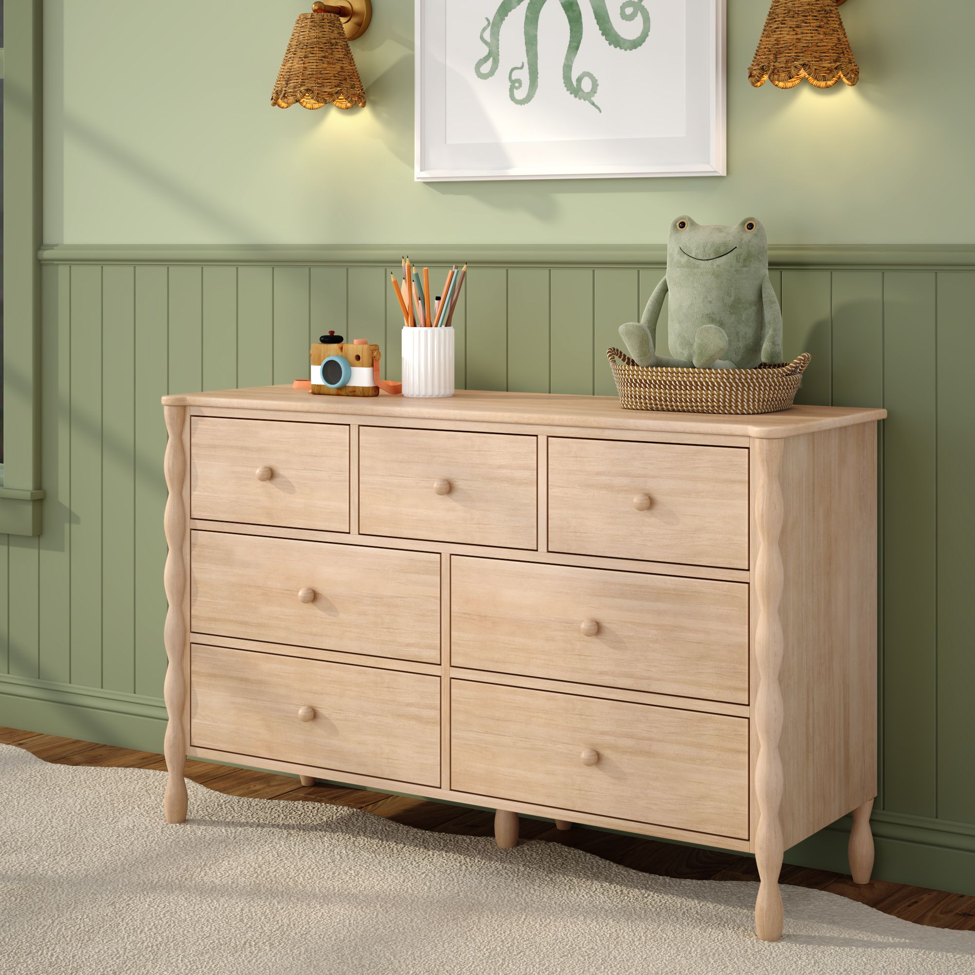 A light-wood Jenny Lind style 7-drawer dresser in a green room. A picture is hanging above the dresser, and small items including a pen cup and stuffed toy are on the dresser.