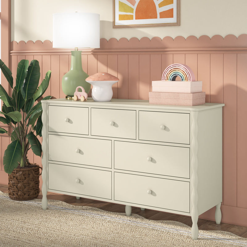 Cream nursery dresser with eight drawers and scalloped legs, topped with green lamp, mushroom nightlight, rainbow stacking toy and pink storage boxes against peach scalloped beadboard wainscoting — boho kids room storage and decor