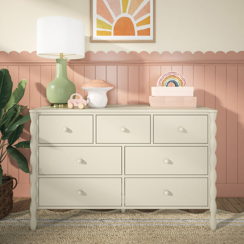 Cream wooden nursery dresser in children's room with green table lamp, mushroom and wooden car toys, pastel rainbow decor, pink scalloped wainscoting and potted plant