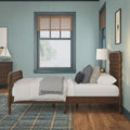 Coastal bedroom interior with wooden spindle bed and tufted headboard, white layered bedding and gray throw, patterned pillows, blue plaid rug, wooden nightstand with sailboat decor and brass lamp, teal walls and bamboo blinds framing an ocean view.