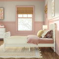 Blush pink children's bedroom with white twin bed, yellow gingham duvet, layered velvet and floral pillows, rattan rug, scalloped wainscoting and Roman shade