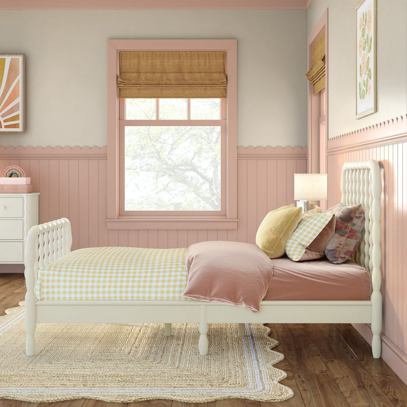 Blush pink children's bedroom with white twin bed, yellow gingham duvet, layered velvet and floral pillows, rattan rug, scalloped wainscoting and Roman shade