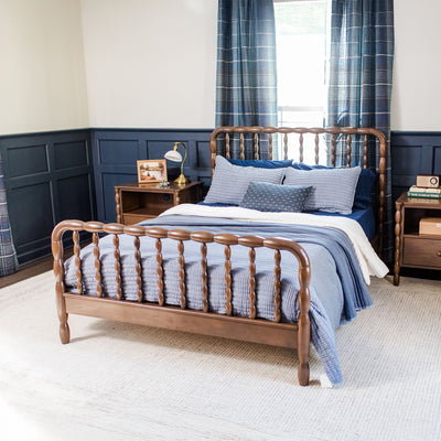 Cozy modern farmhouse bedroom with wooden spindle bed, blue quilted bedding, navy wainscoting, plaid curtains and a nightstand with lamp.