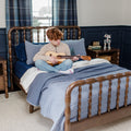Red-haired boy playing ukulele on bed in navy-blue bedroom with wooden spindle bedframe, layered blue bedding and toy robot on nightstand