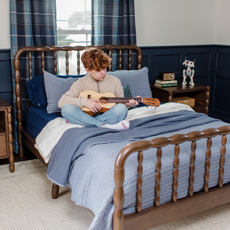 Red-haired boy playing ukulele on bed in navy-blue bedroom with wooden spindle bedframe, layered blue bedding and toy robot on nightstand