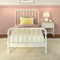 Pastel pink nursery bedroom with white spindle bed, yellow gingham bedding and throw pillow, scalloped wainscoting, white bedside table with lamp and alarm clock