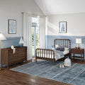 Coastal-themed children's bedroom with wooden spindle bed, blue wainscoting, dark wood dresser, sailboat wall art, plush whale, gray-blue area rug and white curtains