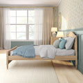 Cozy light-filled bedroom with natural wood single bed, blue throw and pillows, teal wainscoting, floral wallpaper, bedside lamp and large window