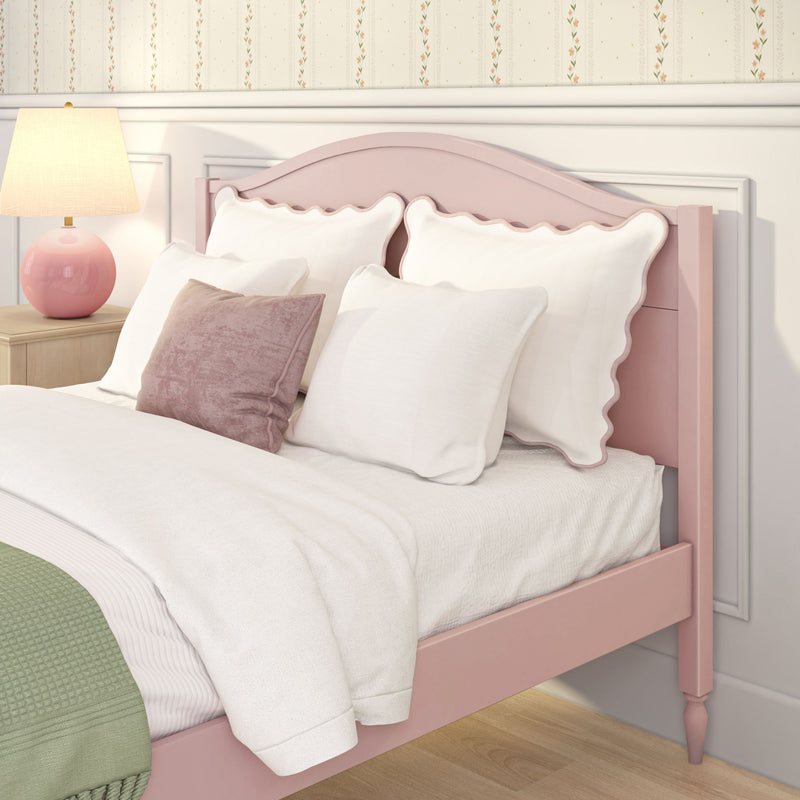 Pastel pink wooden bed with scalloped headboard, white ruffled pillows, pink velvet accent pillow, white comforter and green throw blanket beside a wooden nightstand with pink ceramic lamp in a cozy girls' bedroom