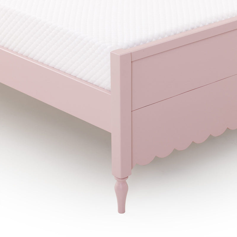 Close-up of pink wooden children's bed frame with scalloped skirt, turned leg and white quilted mattress