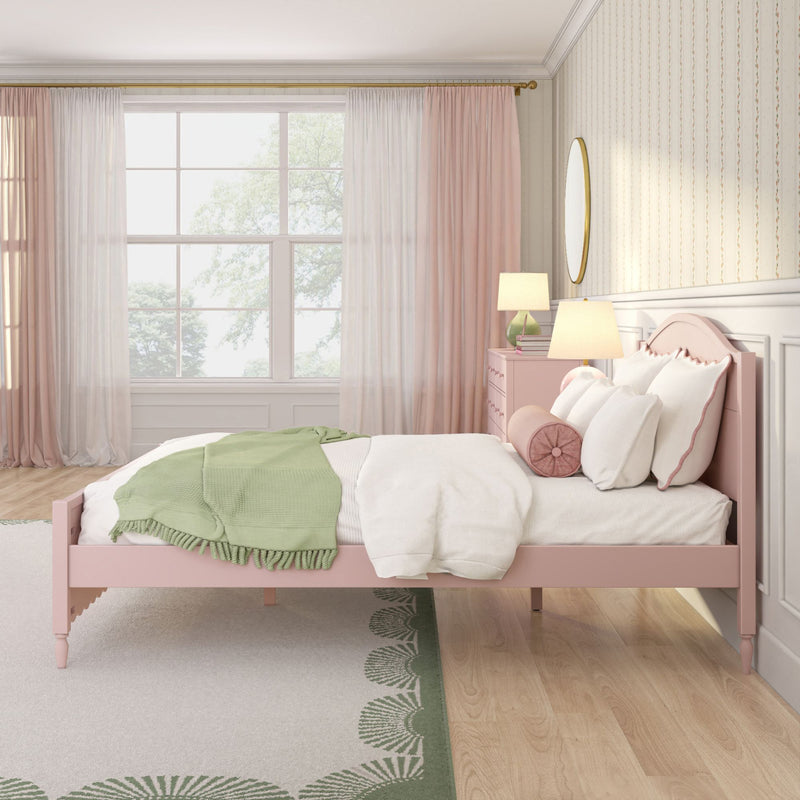 Pastel pink bedroom interior with pink wooden bed, white bedding and green throw blanket, round bolster pillow, pink nightstand with bedside lamps, large window with sheer pink curtains, oval wall mirror and green-accented patterned rug