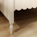 Cream scalloped bed skirt with turned white bed leg on dark hardwood floor, close-up bedroom decor and bed frame detail
