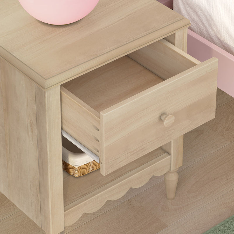 Light wood nightstand bedside table with an open drawer, wicker basket on lower shelf and pink lamp beside bed