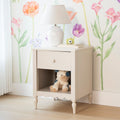 Pastel nursery nightstand with white ceramic lamp, floral watercolor wallpaper, stack of children's books, yellow toy camera and teddy bear